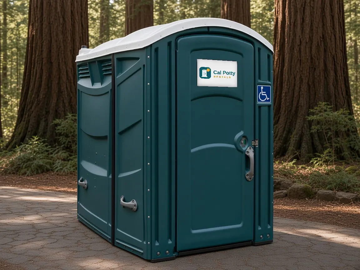 A teal ADA-accessible portable restroom with a wheelchair sign on a paved path under tall redwood trees
